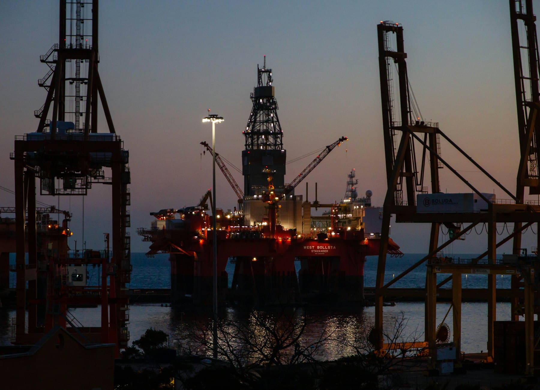 Drilling rig berthed in a harbour among cranes at dusk